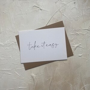 Puede incluir: Una tarjeta blanca con el texto "Take it easy" escrito en cursiva negra. La tarjeta est&aacute; sobre un sobre marr&oacute;n.