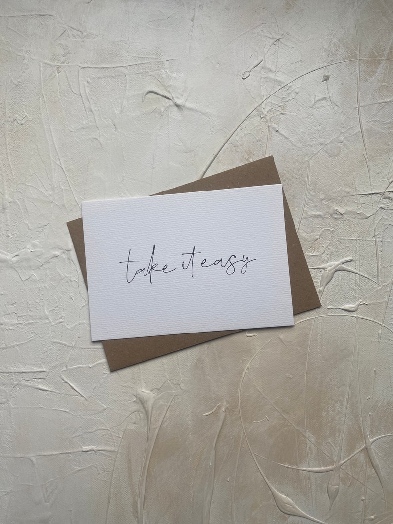 Puede incluir: Una tarjeta blanca con el texto "Take it easy" escrito en cursiva negra. La tarjeta est&aacute; sobre un sobre marr&oacute;n.