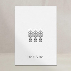 Könnte beinhalten: Eine weiße Karte mit drei schwarz gezeichneten Nussknackersoldaten. Darunter steht in einfacher Schrift "HO HO HO". Das Design ist minimalistisch und die Karte vor einem neutralen Hintergrund platziert.