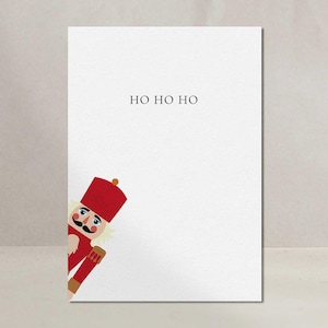 Könnte beinhalten: Eine weiße Grußkarte mit dem Text "HO HO HO" in Schwarz. Ein Cartoon-Nussknacker in roter Uniform und Hut schaut von der linken Seite der Karte hervor. Neutraler Hintergrund.