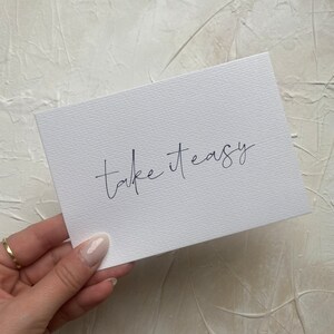 Puede incluir: Tarjeta blanca con el texto "take it easy" escrito en cursiva negra.