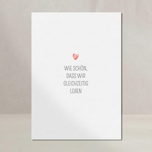 Könnte beinhalten: Eine weiße Karte mit dem deutschen Text "WIE SCHÖN, DASS WIR GLEICHZEITIG LEBEN" und einem kleinen roten Herz. Der Text ist in einer einfachen, serifenlosen Schriftart gehalten. Die Karte wird vor einem neutralen Hintergrund angezeigt.