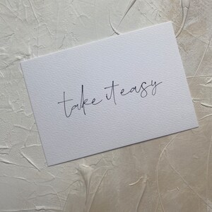 Puede incluir: Una tarjeta blanca con el texto "take it easy" escrito en cursiva negra.
