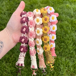 May include: Five crocheted keychains with daisy flower designs in different colors, including burgundy, pink, cream, yellow, and green. Each keychain has a gold metal clasp.