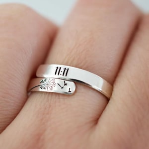 May include: A silver-colored wrap ring with the engraved number "11:11" on the top band. The lower band features a dandelion design with flying birds. The ring is designed to wrap around the finger.