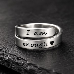 May include: A silver-toned wrap ring featuring a smooth, polished finish. The band is engraved with the black text "I am" on the upper portion and "enough" followed by a small heart symbol on the lower portion of the wrap design.
