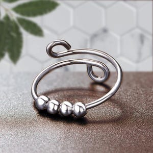 May include: A silver-colored fidget ring with a unique design. The ring features a curved band with four small, round beads and a looped top. The ring is shown on a brown surface.