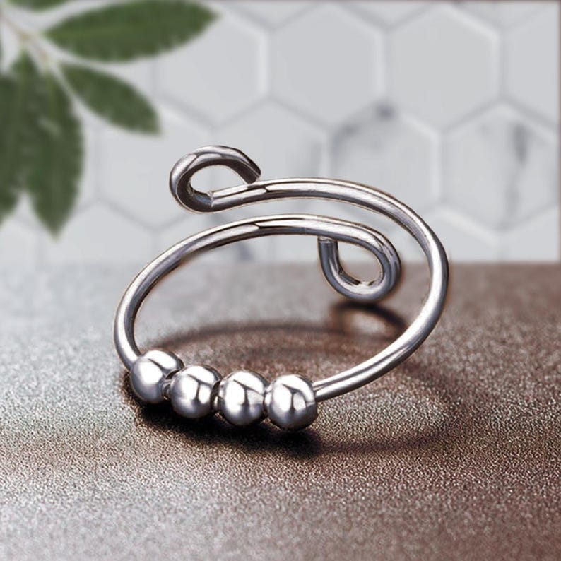 May include: A silver-colored fidget ring with a unique design. The ring features a curved band with four small, round beads and a looped top. The ring is shown on a brown surface.