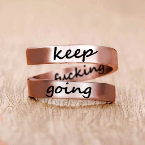 May include: A rose gold-colored ring with the words "keep going" engraved on it. The ring is a double band, with the words "keep" and "going" on separate bands.
