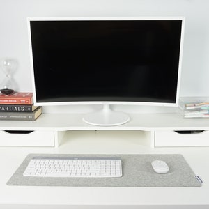 Front view of white desk with light grey (27 x 11 size) wool desk pad on top. White keyboard and mouse on desk mat with black screen in background against wall. The desk mat has enough space for both the keyboard and mouse to be used.