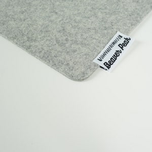 Close up of light grey desk mat corner with Beaver Peak logo on one edge. The logo is made of white cotton with a black embroidered Beaver Peak logo and black stitching to attach it to the desk mat. The logo protrudes from the desk mat edge.