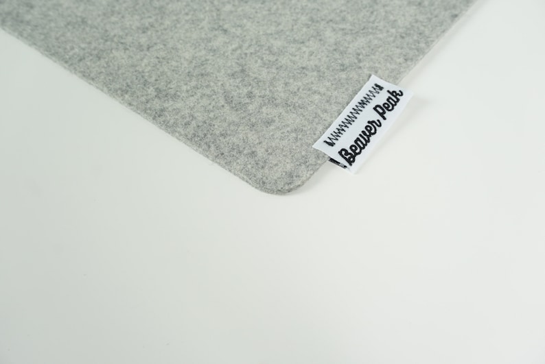 Close up of light grey desk mat corner with Beaver Peak logo on one edge. The logo is made of white cotton with a black embroidered Beaver Peak logo and black stitching to attach it to the desk mat. The logo protrudes from the desk mat edge.