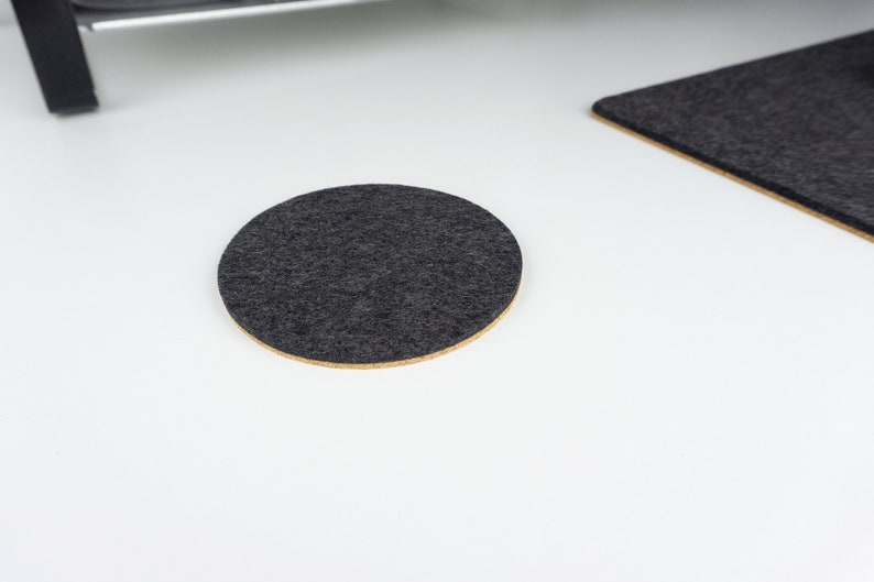 A single circle felt and cork coaster in black felt shown on a white desk surface next to matching black wool felt desk mat.