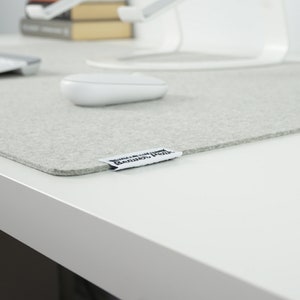 Close up of extra large light grey merino wool desk mat with white keyboard, white mouse, and matching white laptop stand. On white desk with book stack on far end of desk surface.