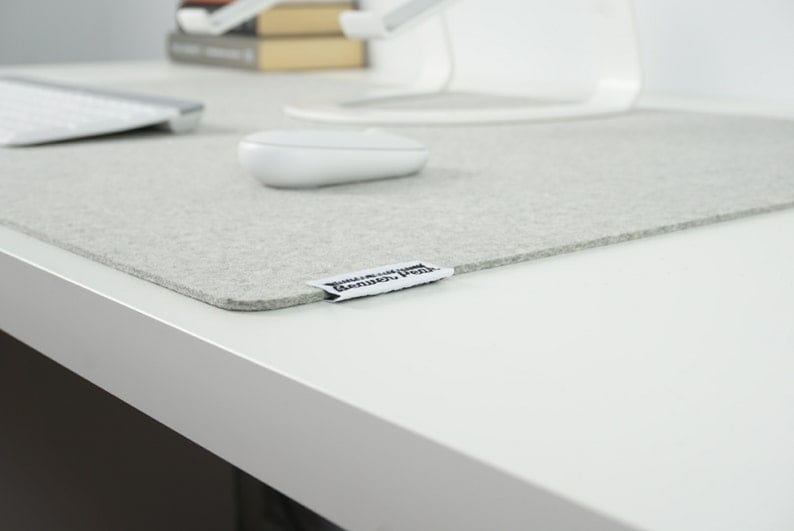 Close up of extra large light grey merino wool desk mat with white keyboard, white mouse, and matching white laptop stand. On white desk with book stack on far end of desk surface.