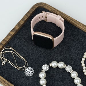 Peut inclure: Un plateau en bois avec un revêtement en feutre gris. Le plateau contient une montre connectée rose et or, un collier en argent avec un pendentif en cristal et un bracelet en perles.