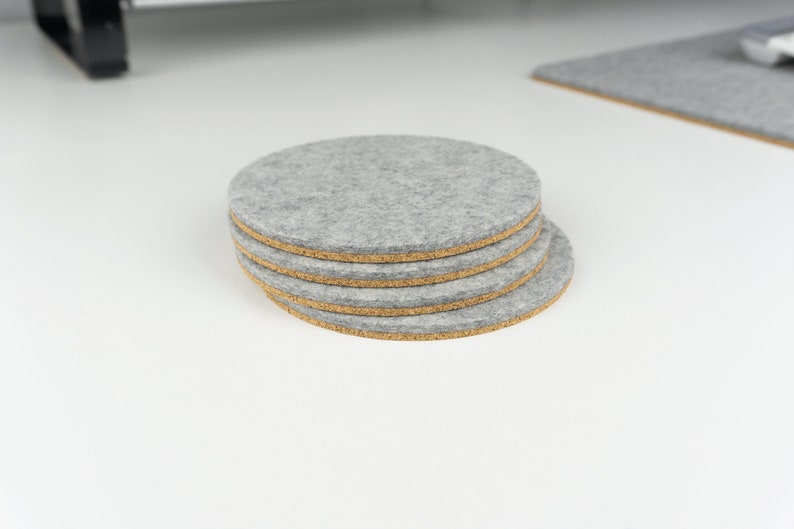 A stack of 4 light grey circular wool felt and cork coasters shown sitting on a white desk next to a matching grey felt desk mat.