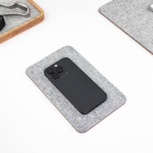 May include: A rectangular light gray felt desk mat with rounded corners, featuring a black smartphone with a triple-lens camera system placed centrally on its surface. The mat provides a soft, textured base for the mobile device.