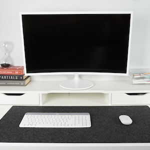 Front view of white desk with black wool desk pad (35 x 17 inch size) on top. White keyboard and mouse on desk mat with black screen in background against wall. Screen sits atop white desk riser lifting it up higher than the desk mat.