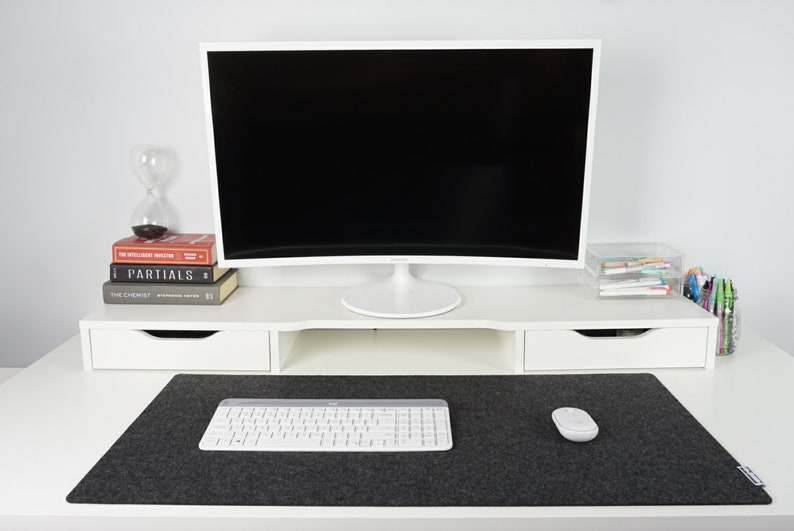 Front view of white desk with black wool desk pad (35 x 17 inch size) on top. White keyboard and mouse on desk mat with black screen in background against wall. Screen sits atop white desk riser lifting it up higher than the desk mat.