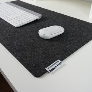 Black (charcoal) merino wool desk mat with  a white keyboard and white mouse. Placed on a white desk setup. Image shows soft texture of the black desk mat with a heather finish against hard white desk.