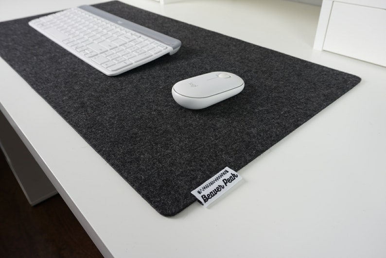 Black (charcoal) merino wool desk mat with  a white keyboard and white mouse. Placed on a white desk setup. Image shows soft texture of the black desk mat with a heather finish against hard white desk.