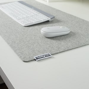 Light grey merino wool desk mat with a white keyboard and a white mouse. Placed on a white desk surface. Image shows soft texture of desk mat against hard white desk.