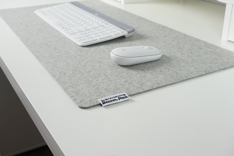 Light grey merino wool desk mat with a white keyboard and a white mouse. Placed on a white desk surface. Image shows soft texture of desk mat against hard white desk.