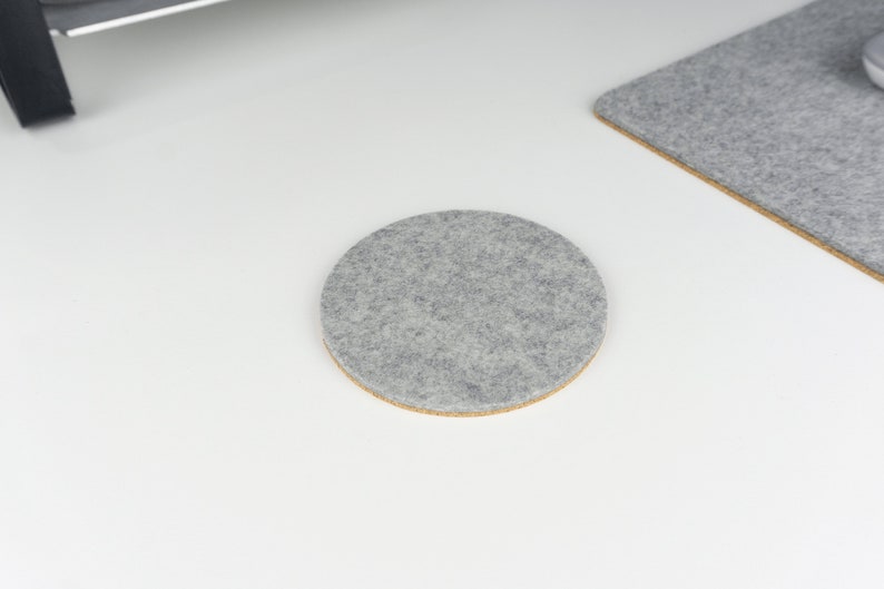 A single round felt and cork coaster in light grey felt shown on a white desk surface next to matching light grey wool felt desk mat.