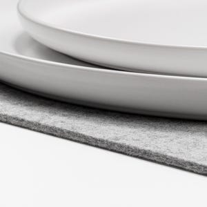 May include: Two stacked white ceramic plates with smooth, rounded edges. The plates are circular in shape and feature a clean, matte finish. They are positioned on a flat, light gray felt mat that has a visible, slightly textured surface.