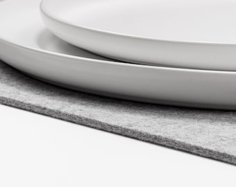 Puede incluir: Dos platos de cerámica blanca apilados con bordes lisos y redondeados. Los platos son de forma circular y presentan un acabado mate limpio. Están colocados sobre una alfombrilla de fieltro gris claro con una superficie visible y texturizada.