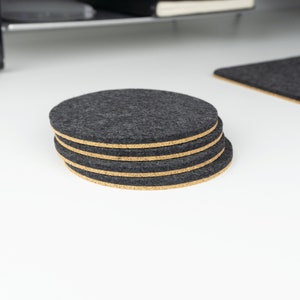 A stack of 4 black circular wool felt and cork coasters shown sitting on a white desk next to a matching felt desk mat.