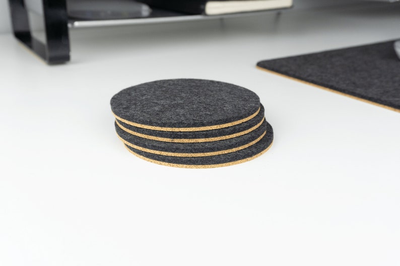 A stack of 4 black circular wool felt and cork coasters shown sitting on a white desk next to a matching felt desk mat.