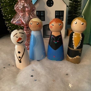 May include: A set of five wooden peg dolls, painted to resemble characters from the movie Frozen. The dolls include Elsa, Anna, Kristoff, Olaf, and a snowman. The dolls are standing on a white surface with silver glitter.