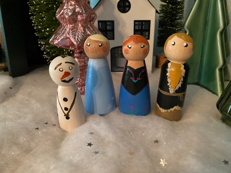 May include: A set of five wooden peg dolls, painted to resemble characters from the movie Frozen. The dolls include Elsa, Anna, Kristoff, Olaf, and a snowman. The dolls are standing on a white surface with silver glitter.