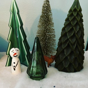May include: A collection of four decorative Christmas trees. The trees are made of different materials, including green ceramic, gold glitter, and green felt. A small wooden snowman figurine is in front of the trees.