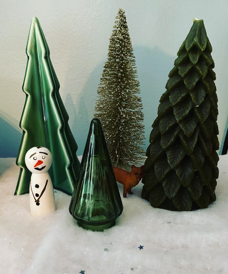 May include: A collection of four decorative Christmas trees. The trees are made of different materials, including green ceramic, gold glitter, and green felt. A small wooden snowman figurine is in front of the trees.