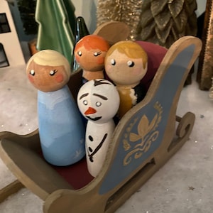 May include: Four wooden peg dolls in a blue and gold sleigh. The dolls are dressed as characters from the movie Frozen. The dolls are a snowman, a girl with blonde hair, a girl with red hair, and a boy with blonde hair.