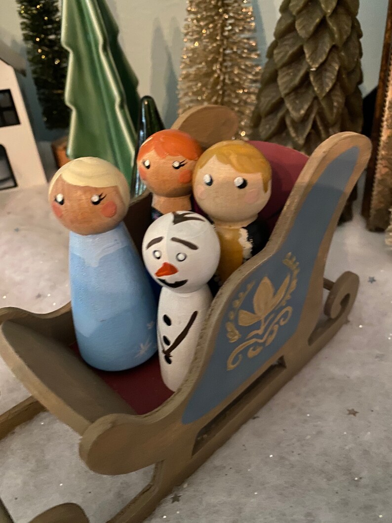 May include: Four wooden peg dolls in a blue and gold sleigh. The dolls are dressed as characters from the movie Frozen. The dolls are a snowman, a girl with blonde hair, a girl with red hair, and a boy with blonde hair.