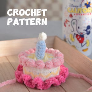 May include: A crocheted cake with pink and white frosting, a blue candle, and the text "CROCHET PATTERN". The cake sits on a wooden tray, with a Disney California Adventure mug in the background.