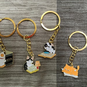 May include: Four gold keychains with different cat-themed charms. One charm features a cat reading a book, another a cat with a magnifying glass, another a cat with a book, and the last a cat sleeping on a book. The charms are attached to gold key rings.