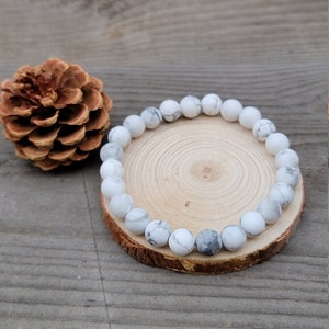 May include: White and gray beaded bracelet with a natural stone appearance. The bracelet is displayed on a small, round wooden slice. A large pine cone is next to the bracelet. The background is a wooden surface.