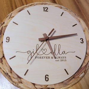Könnte beinhalten: Eine runde Holzuhr mit hellem Holzgesicht und dunkelbraunen Zeigern und Ziffern. Das Zifferblatt zeigt die Namen "gil & ella" mit einem Herzsymbol und dem Text "FOREVER & ALWAYS est 2015". Die Uhr steht auf einem gewebten, kreisförmigen Sockel.