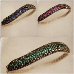 May include: Three silver bangle bracelets featuring a row of colourful gemstones and a row of black marcasite stones. The bracelets are displayed on a white background.