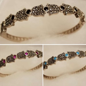 Pode incluir: Um bracelete de prata com um design delicado que apresenta uma fila de engastes em forma de marquesa com pedras preciosas pretas, rosas e azuis. O bracelete é feito de prata de lei e é adornado com marcasite.