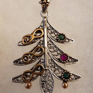 May include: A silver filigree pendant in the shape of a tree with gold accents and green and pink gemstones. The pendant is attached to a silver chain.