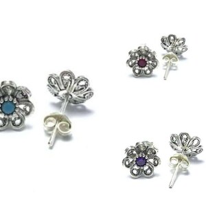May include: Three pairs of silver flower-shaped earrings with different colored gemstones in the center. The earrings have a filigree design and are on a white background.