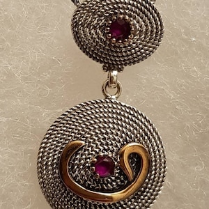 Könnte beinhalten: Eine Halskette aus Silber und Gold mit einem Anhänger mit zwei kreisförmigen Designs. Der Anhänger hat ein gewebtes Muster und zwei violette Edelsteine.