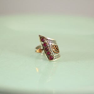 May include: A silver ring with a gold filigree design and red gemstones. The ring is angled and has a unique shape.