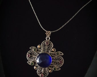 Peut inclure: Un collier avec un pendentif en filigrane argenté orné d'une grande pierre précieuse bleue entourée de petites pierres précieuses vertes et rouges.
